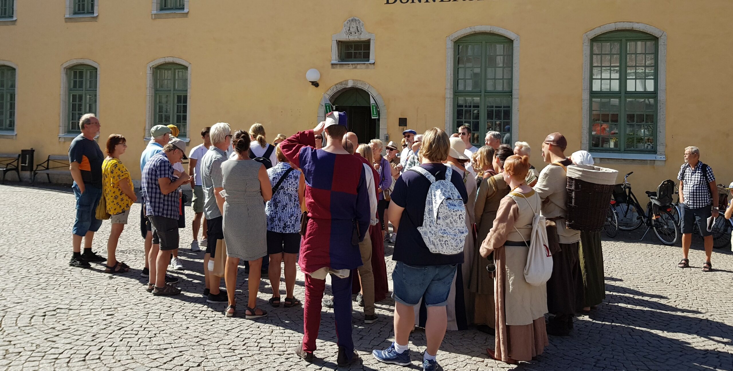 Människor lyssnar på guide som berättar om Visby utanför Turistbyrån på Donners plats