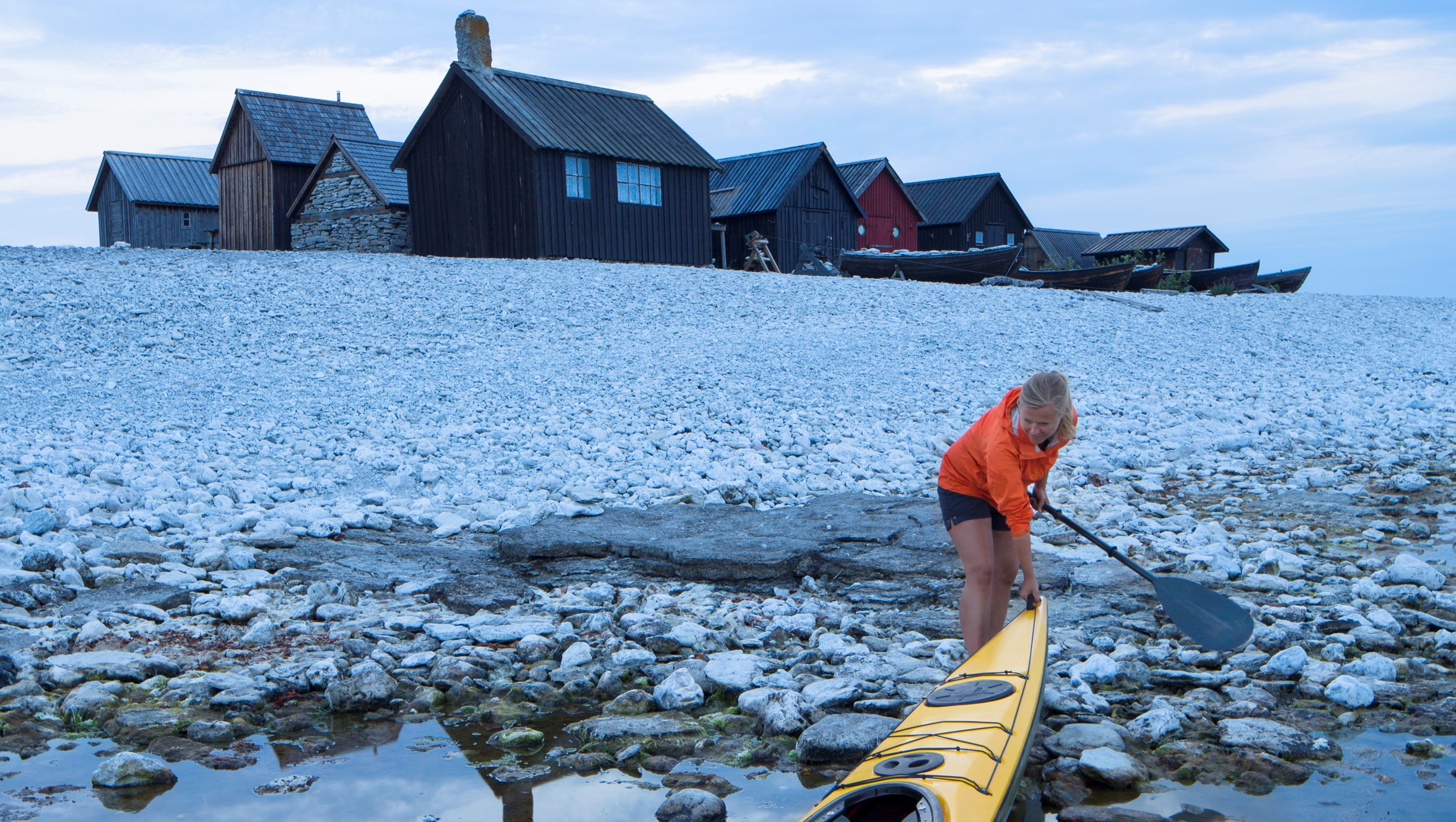 Kvinna drar upp kajak på stenstrand framför ett fiskeläge.